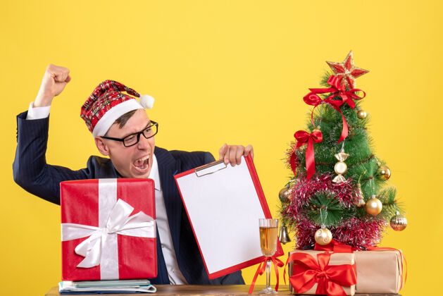 礼物商务人士的正面图显示 他坐在圣诞树旁的桌子旁 展示着胜利的姿态 黄色的礼物桌子胜利圣诞树