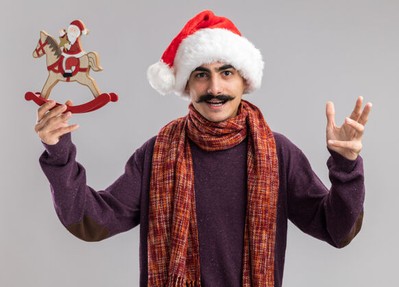 圣诞快乐年轻的胡子男人戴着圣诞老人帽 脖子上围着暖和的围巾 手里拿着圣诞玩具 兴高采烈地看着相机 举起胳膊站在白色的背景上周围胡子帽子
