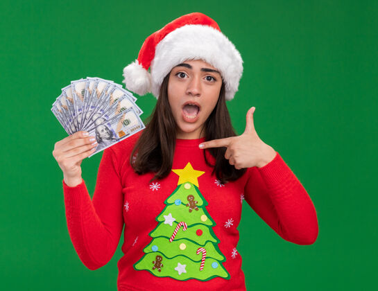 女孩焦急的年轻白人女孩戴着圣诞老人的帽子 拿着钱指着绿色背景上的复制空间圣诞老人年轻新年