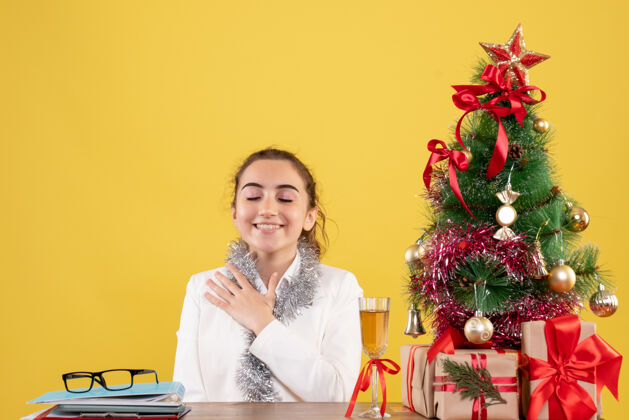女医生正面图女医生围坐在圣诞礼物和黄色背景上微笑的圣诞树旁周围微笑礼物