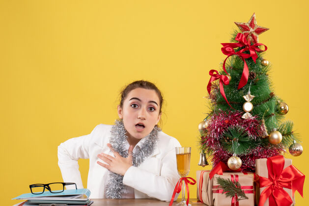 背景正面图女医生围坐在黄色背景上的圣诞礼物和圣诞树旁礼物人健康