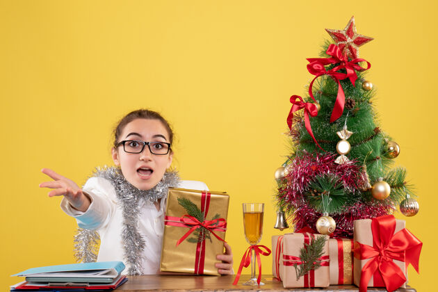 健康前视图：女医生坐在桌子后面 拿着圣诞礼物 黄色背景上的树坐背景前面