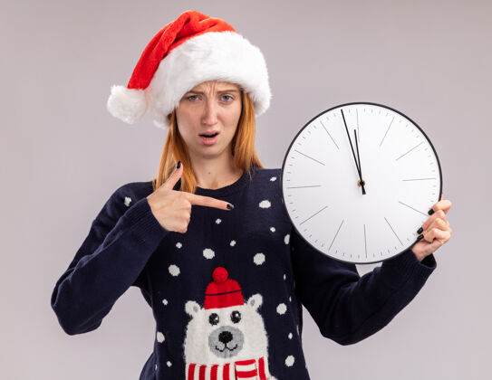 钟迷茫的年轻漂亮女孩戴着圣诞帽 指着隔离在白色背景上的挂钟举行墙女孩