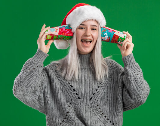 耳朵快乐的年轻金发女人 穿着冬衣 戴着圣诞帽 耳朵上戴着五颜六色的纸杯 站在绿色的背景上 欢快地笑着圣诞老人微笑圣诞