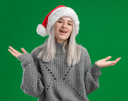 金发年轻的金发女郎穿着冬衣 戴着圣诞帽 站在绿色的背景下 开心而积极地微笑着看着镜头快乐帽子站着