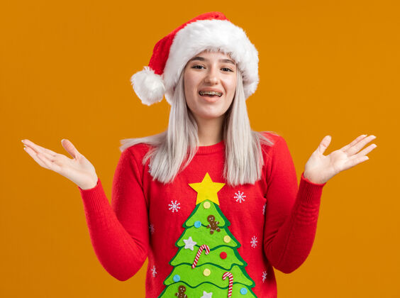 毛衣年轻的金发女郎穿着圣诞毛衣 戴着圣诞帽 开心地看着镜头 积极地张开双臂站在橙色背景的一边年轻手臂伸展