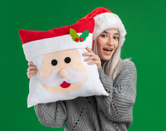 抱着快乐的金发女郎 穿着冬衣 戴着圣诞帽 抱着圣诞枕头 面带微笑地看着相机 站在绿色的背景下帽子年轻圣诞老人