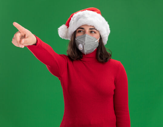站着穿着红色毛衣 戴着圣诞老人帽 戴着护面面具的年轻女孩一边看着 一边开心地用食指指着绿墙上站着的东西面部指向圣诞老人
