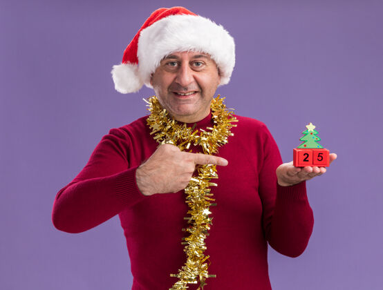 手指快乐的中年男人戴着圣诞老人帽子 脖子上戴着金属丝 手里拿着玩具方块 上面写着“25岁”的日期 用食指指着他们 站在紫色的背景上微笑着站着指数脖子