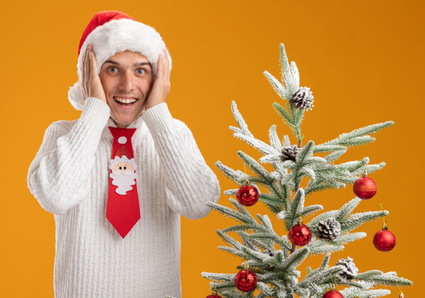 靠近令人印象深刻的年轻帅哥戴着圣诞帽 打着圣诞老人的领带站在装饰好的圣诞树旁 手放在头上 隔离在橙色的墙上印象深刻帽子年轻