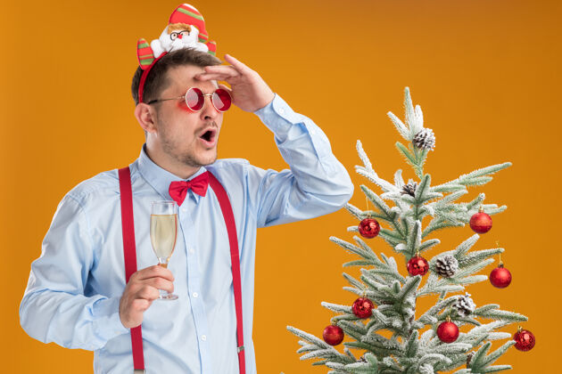 蝴蝶结戴着吊带领结的年轻人站在圣诞树旁 戴着圣诞老人和红眼镜 手里拿着一杯香槟 远远地看着橙色的墙上远头圣诞老人