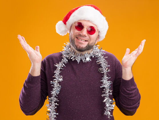 圣诞老人微笑的中年男子戴着圣诞帽 脖子上戴着金箔花环 戴着眼镜 两手空空的 孤立在橙色的墙上金属丝脖子展示