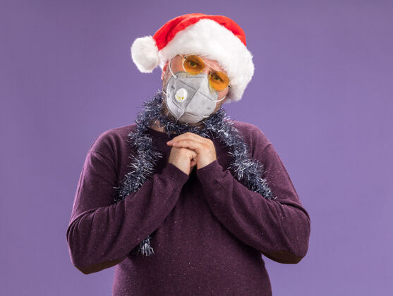 花环困惑的中年男子戴着圣诞老人帽 戴着防护面具 脖子上戴着金属丝花环 戴着眼镜 手放在一起 在紫色的墙上与世隔绝帽子脖子圣诞节