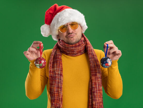 不高兴一个穿着黄色高领毛衣 戴着圣诞帽 戴着眼镜 拿着圣诞树玩具的年轻人站在绿色的墙上 看上去既困惑又不高兴玩具困惑穿着