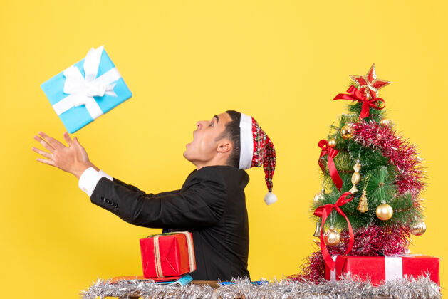 抓住正面图：一个戴着圣诞帽的男人坐在桌子旁 试图抓住他现在的圣诞树和礼物坐着帽子人