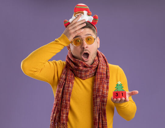 显示穿着黄色高领毛衣 戴着暖和的围巾和眼镜的年轻人戴着滑稽的帽沿 头上戴着圣诞老人 他站在紫色的墙上 展示着印有新年日期的玩具立方体年轻高领毛衣围巾