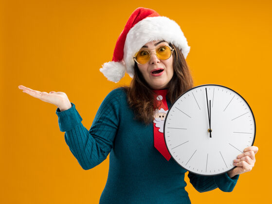 橙色戴着太阳眼镜 戴着圣诞帽 系着圣诞领带的成年白种女人拿着闹钟 手一直开着 手被隔离在橙色的背景上 还有复印空间眼镜打开手