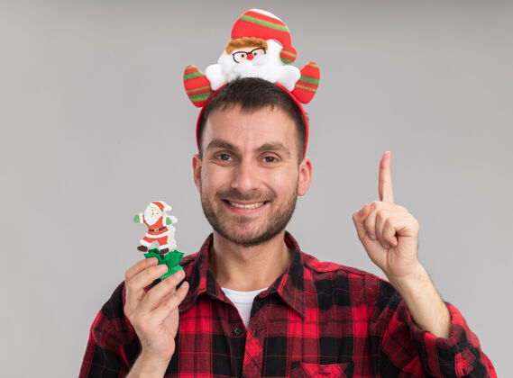 指着微笑着的白人年轻人戴着圣诞老人的头带 手里拿着雪人圣诞玩具 看着孤立地指向白色背景的摄像机抱着雪人圣诞老人