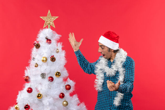 帽子一个快乐的年轻人戴着圣诞老人的帽子 穿着一件蓝色的衬衫 看着圣诞树 红得惊人圣诞树圣诞老人脱光衣服