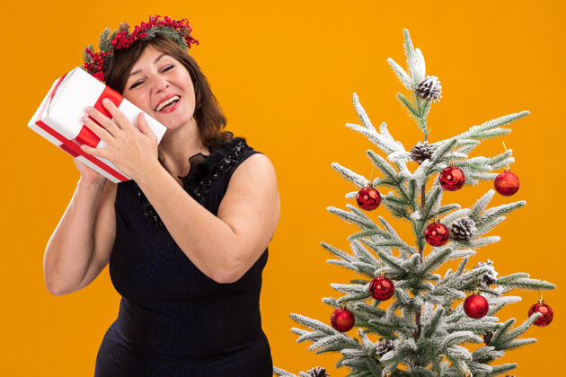 眼睛快乐的中年妇女头戴圣诞花环 脖子上戴着金箔花环 站在装饰好的圣诞树旁包装年佩戴