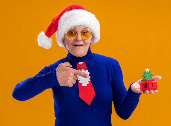抱着戴着太阳眼镜 戴着圣诞帽 系着圣诞领带 面带微笑的老妇人拿着圣诞树的装饰物 这些装饰物被隔离在橙色背景上 并留有复制空间橘子领带老人