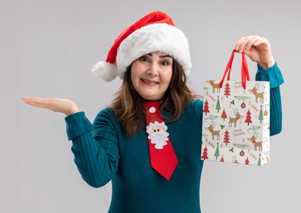 领带微笑的成年白人妇女 戴着圣诞帽 打着圣诞领带 手里拿着一个纸礼盒 在白色背景上 手一直张开 并留有复印空间帽子圣诞老人微笑