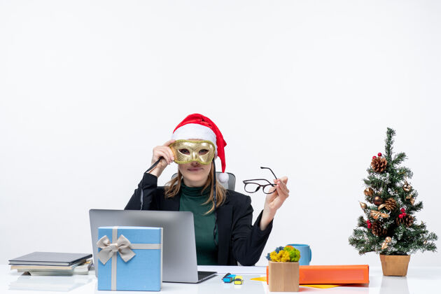 年轻人戴着圣诞老人帽 戴着眼镜 戴着面具的年轻女子坐在桌子旁 手里拿着圣诞树和礼物桌子工作场所年轻女人