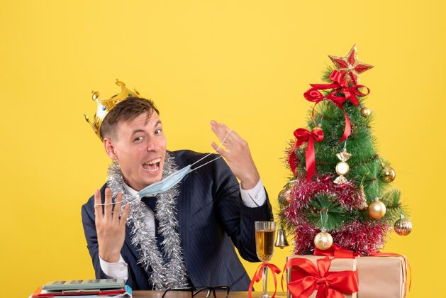 礼物正面图：快乐的商人手持面具坐在圣诞树旁的桌子旁 黄色的礼物礼物圣诞节庆祝