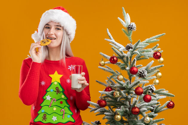 女人穿着圣诞毛衣 戴着圣诞帽 拿着一杯牛奶 吃着饼干的金发女郎快乐而积极橙色圣诞老人积极