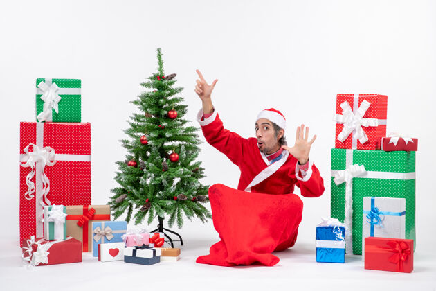 人年轻的成年人打扮成圣诞老人 拿着礼物和装饰好的圣诞树坐在地上指着上面的五棵打扮年轻人五