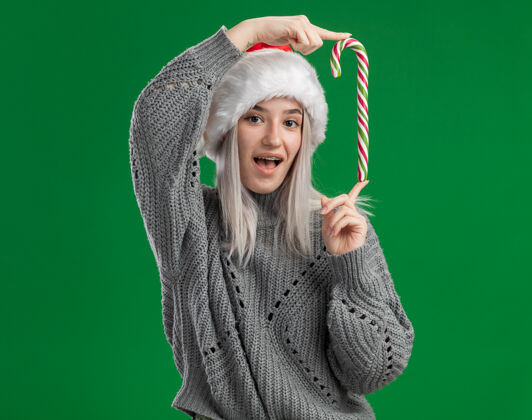 拿着年轻的金发女郎穿着冬衣 戴着圣诞帽 手里拿着糖果手杖 开心地看着站在绿色背景下的摄像机圣诞老人手杖金发