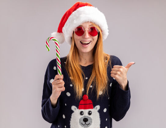 年轻快乐的年轻漂亮的女孩戴着圣诞帽 戴着圣诞眼镜 拿着圣诞糖果 在白色的墙上孤立地竖起大拇指糖果拇指穿着