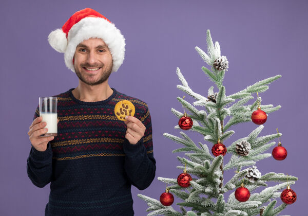 年轻人微笑着的白人年轻人戴着圣诞帽站在装饰好的圣诞树旁 手里拿着一杯牛奶和饼干 看着隔离在紫色背景上的摄像机相机饼干牛奶