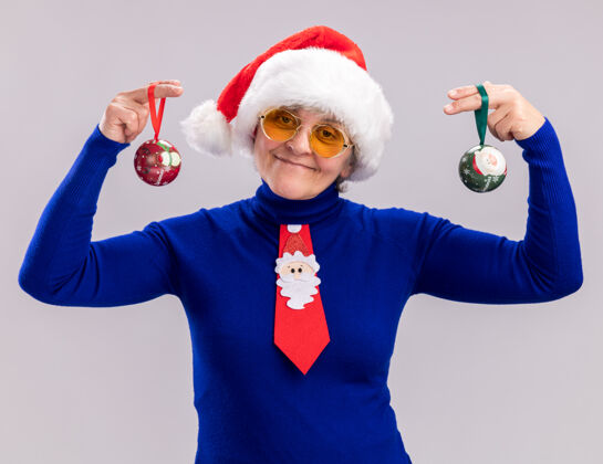 玻璃戴着太阳眼镜 戴着圣诞帽 系着圣诞领带 拿着玻璃球的老妇人高兴地说 这些装饰物是在白色背景上孤立的 还有复制空间装饰品抱着高兴