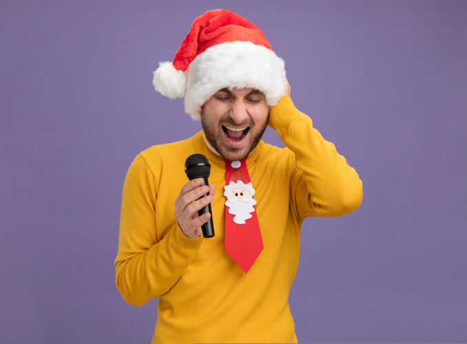 眼睛紧张的白人年轻人戴着圣诞帽打着领带拿着麦克风手放在头上尖叫着闭着眼睛隔离在紫色的背景和复制空间尖叫年轻麦克风