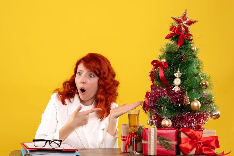 情感女工人坐在桌子后面 手里拿着圣诞礼物和黄色的圣诞树圣诞女黄色