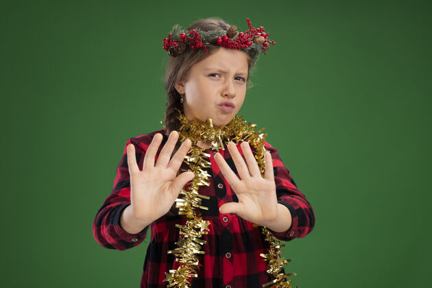 制造戴着圣诞花圈的小女孩穿着格子裙 脖子上戴着金属丝 看着摄像机 担心地做着防御手势 双手站在绿色背景上周围手势担心
