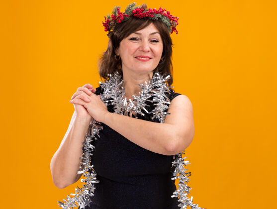 微笑微笑的中年妇女头戴圣诞花环 脖子上戴着金属丝花环 双手合十 看着橙色背景上的相机 还有复制空间脖子周围头