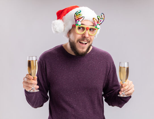 圣诞老人快乐的年轻人穿着紫色毛衣 戴着圣诞帽 戴着搞笑的眼镜 手里拿着两杯香槟 兴高采烈地微笑着看着镜头 新年假期的概念站在白色的背景上微笑眼镜帽子