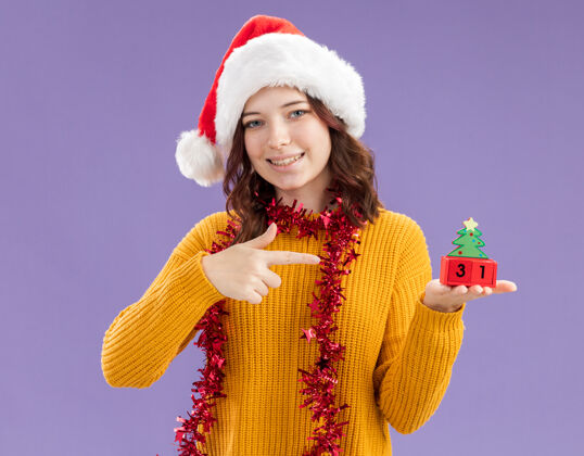 斯拉夫人微笑着的年轻斯拉夫女孩 戴着圣诞帽 脖子上戴着花环 拿着并指着紫色背景上孤立的圣诞树装饰 还有复制空间抱着年轻指着