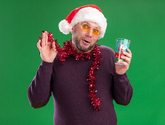 圣诞节令人印象深刻的中年男子戴着圣诞帽 脖子上戴着金属丝花环 戴着眼镜 手里拿着塑料圣诞杯 手里拿着金属丝花环 看着隔离在绿色背景上的相机塑料脖子举行