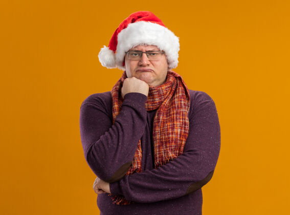 脖子眉头紧锁的成年男子戴着眼镜 戴着圣诞帽 脖子上围着围巾 拳头放在下巴上 看着橙色背景上孤立的摄像机眼镜下巴帽子