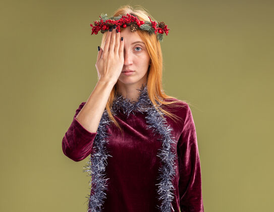 花环年轻漂亮的女孩穿着红色的裙子 脖子上戴着花环和花环 手被隔离在橄榄绿的墙上 遮住了眼睛封面礼服橄榄