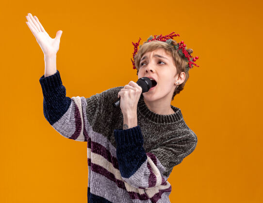 头年轻漂亮的女孩戴着圣诞花环 拿着麦克风 近在嘴边举手 抬头唱歌 隔离在橙色的墙上 有复制空间穿着近漂亮