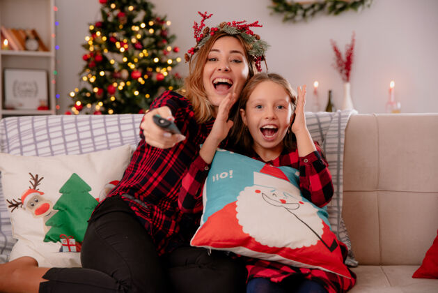 看带着冬青花环的快乐妈妈拿着电视遥控器 女儿坐在沙发上 在家里享受圣诞节时光举行冬青母亲