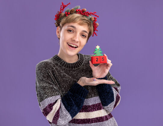 花环快乐的年轻漂亮女孩戴着圣诞花环 手里拿着圣诞树玩具 日期指向它 隔离在紫色的墙上 有复制空间头日期指向