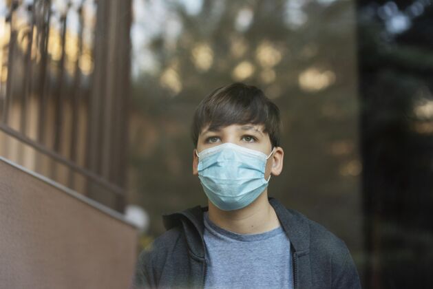 男孩戴着医用口罩的男孩看着窗外的窗户呆在家里流感健康
