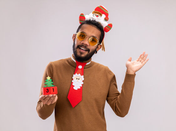 微笑一个穿着棕色毛衣 头戴圣诞老人戒指 打着有趣的红色领带的非裔美国人拿着新年约会的玩具立方体 站在白色背景下 面带微笑地看着相机日期脸非洲