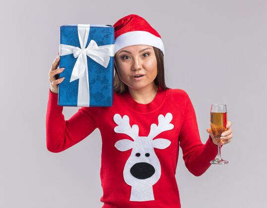 杯子看着镜头年轻的亚洲女孩戴着圣诞帽拿着毛衣拿着礼品盒一杯香槟隔离在白色背景上帽子年轻人香槟