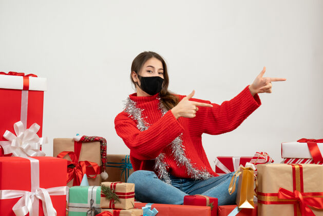 圣诞节穿着红色毛衣 戴着黑色口罩的年轻女孩手指着什么东西坐在白色的礼物上黑色礼物一些东西